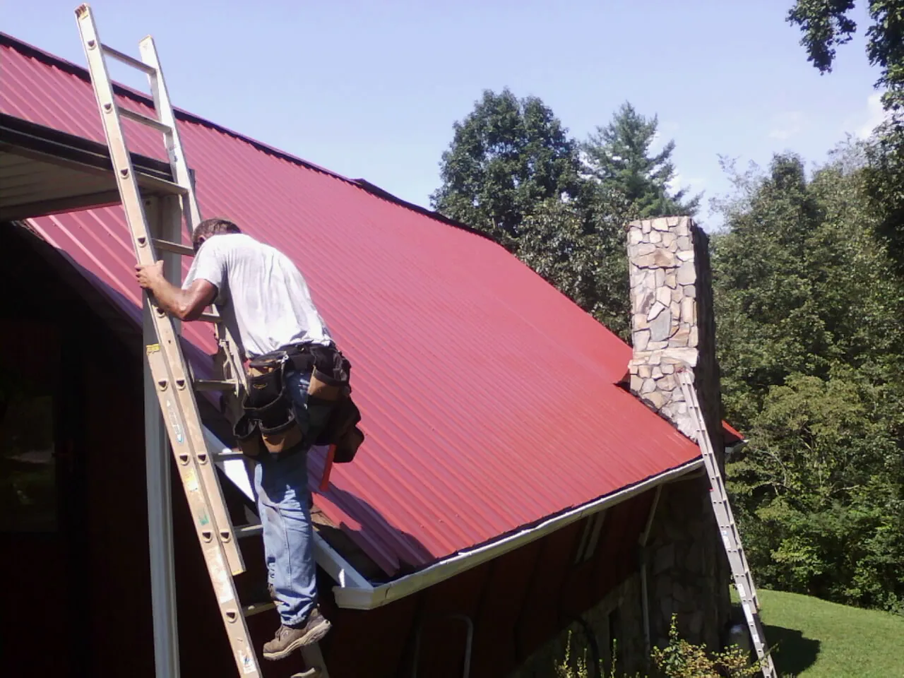 Roofer on a ladder beside a red metal roof and stone chimney — Soffit & Fascia Repair by Roofing Brothers in Rowesville